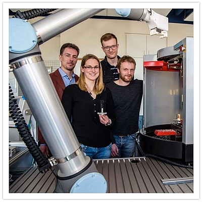 vier Personen im Labor im Vordergrund Teile von Roboter und Fräsmaschine