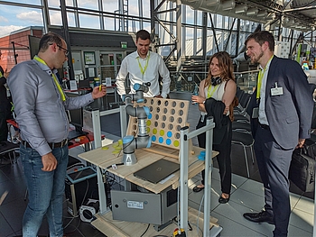 Vier Personen stehen in einer Halle um einen Versuchsstand mit Roboter und einem übergroßen Spiel, wo zweifarbige Scheiben durch den Roboter eingesteckt werden können